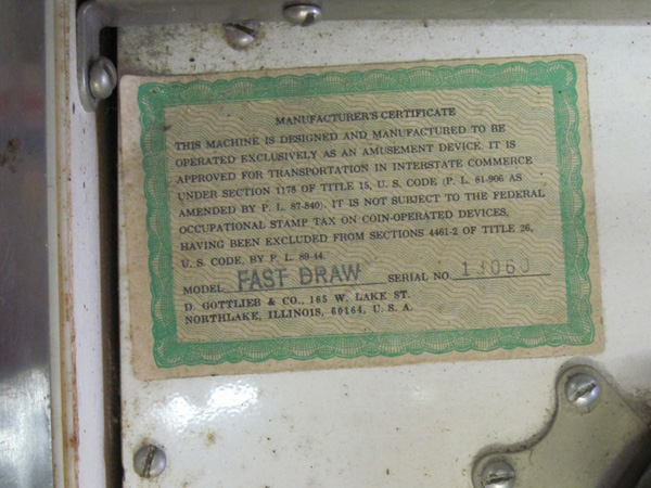 A closeup of the "Manufacturer's Certificate" above the upper left playfield. The background is a green, curlicued certificate frame and resembles currency or an old stock note. It reads, in all caps, "This machine is designed and manufactured to be operated exclusively as an amusement device. It is approved for transportation in interstate commerce under section 1178 of Title 15, U.S. Code (P.L. 81-906 as amended by P.L. 87-840). It is not subject to the Federal Occupational Stamp Tax on Coin-Operated Devices, having been excluded from sections 4461-2 of Title 26, U.S. Code, by P.L. 89-44." The model is stamped "Fast Draw" and the serial number is 13060. The address on the bottom reads "D. Gottlieb & Co., 185 Lake St. / Northlake, Illinois, U.S.A."  